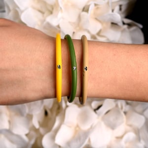 May include: Three colorful bangles: yellow, green, and tan. Each bangle has a small, round, silver-colored embellishment. The bangles are worn on a wrist, with a white floral background.