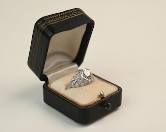 Puede incluir: Un anillo de diamantes en un joyero negro de estilo vintage. El anillo presenta un diamante central grande de talla redonda, rodeado de diamantes más pequeños. El joyero tiene un interior color crema y un ribete dorado.