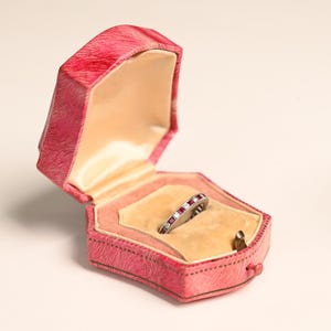 May include: A silver ring with alternating red and clear stones rests inside a pink, velvet-lined jewelry box. The box is open, revealing a peach-colored interior and a gold-colored clasp. The box has a textured exterior.