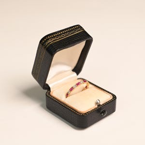 May include: A gold ring with a series of red gemstones and small diamonds is displayed inside a black jewelry box. The box is open, revealing a cream-colored interior. The box has a decorative gold pattern around the edges.