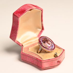 Può includere: Un anello d'oro con una grande gemma rosa ovale e una fila di gemme rosse più piccole, incastonato in un portagioie di velluto rosa. Il portagioie è aperto, rivelando un interno color crema.