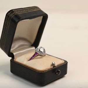 May include: A heart-shaped diamond ring with a halo of smaller diamonds, set in a silver band with a row of pink gemstones. The ring is displayed in an open black leatherette jewelry box with a cream-colored interior.