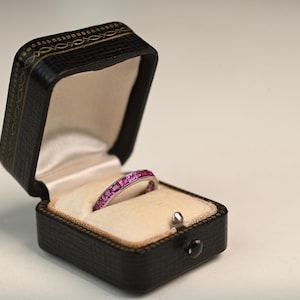 May include: A silver ring with a band of pink gemstones rests inside an open black jewelry box. The box has a cream-colored interior and a decorative gold trim around the edges. The ring is the focal point.
