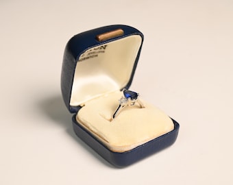 May include: A silver ring with a blue, heart-shaped gemstone and two smaller white gemstones, displayed in an open, navy blue jewelry box with a cream-colored interior. The box is rectangular with a rounded top.