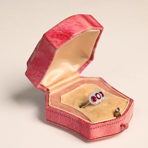 Op de afbeelding: Een vintage ring met drie ovale rode edelstenen en kleine witte stenen gezet in een gouden band, gepresenteerd in een open roze juwelendoos met een gouden interieur.
