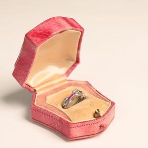 Puede incluir: Un anillo de compromiso plateado con una piedra central y piedras de acento más pequeñas descansa en un joyero rosa hexagonal. El joyero tiene un interior color melocotón y un cierre. Una joya clásica.