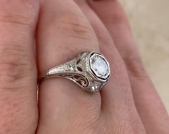 May include: A vintage-style ring with a large, round, clear gemstone set in a filigree design. The ring is made of a light-colored metal, possibly silver or white gold, and features intricate openwork details. The ring is a classic piece of jewelry.