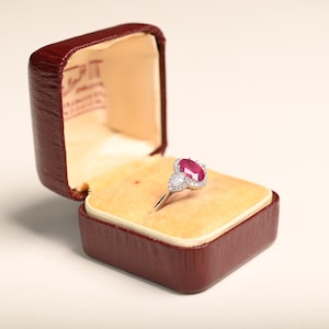 May include: A ruby and diamond ring displayed in an open, vintage, burgundy leather ring box. The ring features a large, oval-cut ruby center stone, surrounded by smaller diamonds. The box has a cream-colored interior.