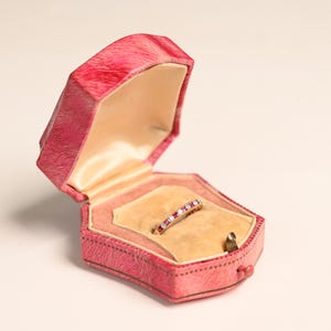 Op de afbeelding: Een vintage roze sieradendoosje met een goudkleurige ring met rode edelstenen. De ring is gezet in een gouden band. De doos is open en onthult een crèmekleurig interieur.