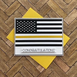 May include: A greeting card with a black and white American flag design, featuring a yellow stripe. The card includes the word "CONGRATULATIONS" on a white banner. It is placed on a yellow envelope, set against a wooden background.