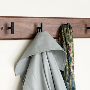 May include: A wooden coat rack with four black metal hooks mounted on a wall. A light grey coat and a patterned scarf hang from the hooks. The wood is a dark brown colour.