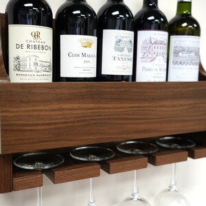 May include: A wall-mounted wooden wine rack with a shelf for storing bottles and a lower rack for hanging wine glasses. The rack is made of dark brown wood and has a natural wood grain finish. Five bottles of wine are displayed on the shelf, and four wine glasses are hanging from the lower rack.