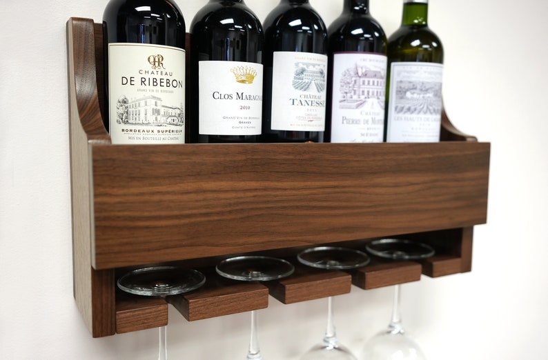 May include: A wall-mounted wooden wine rack with a shelf for storing bottles and a lower rack for hanging wine glasses. The rack is made of dark brown wood and has a natural wood grain finish. Five bottles of wine are displayed on the shelf, and four wine glasses are hanging from the lower rack.