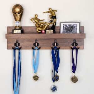 May include: A wooden wall-mounted shelf with four hooks for hanging medals. The shelf has a brown finish and is decorated with a soccer trophy, a framed photo of a soccer team, and a framed photo of two soccer players. The shelf is mounted on a white wall.