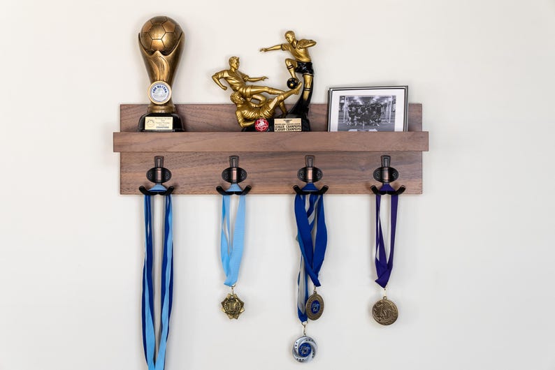 May include: A wooden wall-mounted shelf with four hooks for hanging medals. The shelf has a brown finish and is decorated with a soccer trophy, a framed photo of a soccer team, and a framed photo of two soccer players. The shelf is mounted on a white wall.