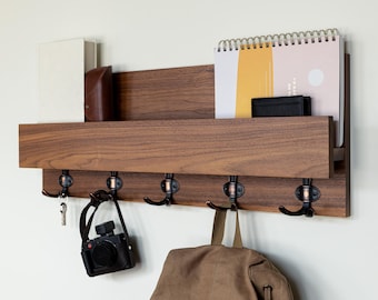 May include: A wooden wall mounted shelf with five hooks and a small shelf on top. The shelf is holding a book, a wallet, and a spiral bound notebook. A camera and a backpack are hanging from the hooks.