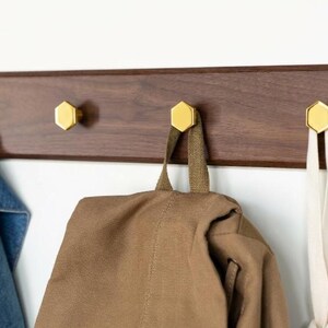 May include: A dark wooden coat rack with four brass hexagonal hooks. A denim jacket, a brown backpack, and a white tote bag hang from the hooks. The rack is mounted on a white wall. The hooks are gold-coloured.