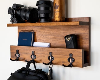 May include: A wooden wall-mounted shelf with hooks and a small shelf for storage. The shelf is made of dark wood and has a natural finish. It features four hooks for hanging items and a small shelf for storing small items. A camera, a notebook, a small box, and a backpack are on the shelf.