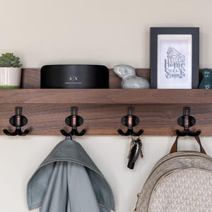 May include: A wooden wall-mounted shelf with four hooks and a small shelf. The shelf is decorated with a potted succulent, a small speaker, a ceramic rabbit figurine, and a framed print that says "Home Sweet Home".