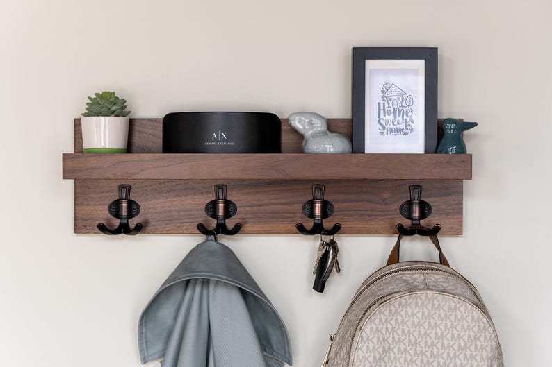 May include: A wooden wall-mounted shelf with four hooks and a small shelf. The shelf is decorated with a potted succulent, a small speaker, a ceramic rabbit figurine, and a framed print that says "Home Sweet Home".