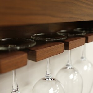 May include: A wooden wine glass rack with four wine glasses hanging from it. The rack is mounted on a wall and has a dark wood finish.