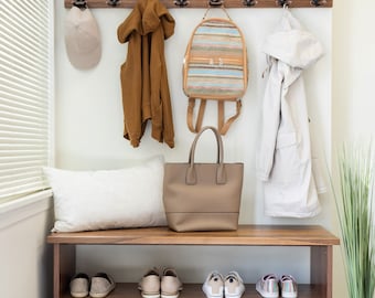 May include: A wooden entryway bench with a shelf for shoes and a top shelf for storage. The bench has a white cushion and a brown tote bag, a backpack, a jacket, and a hat hanging on hooks above it. There is a basketball and a wicker basket on the floor.