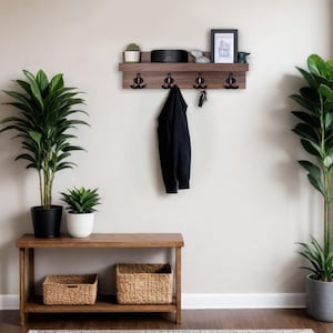 Peut inclure: Une étagère murale en bois avec quatre crochets et une petite étagère sur le dessus. L'étagère est brune et les crochets sont noirs. Il y a un haut-parleur noir, une petite plante et un cadre photo sur l'étagère supérieure. Une veste noire est accrochée à l'un des crochets.