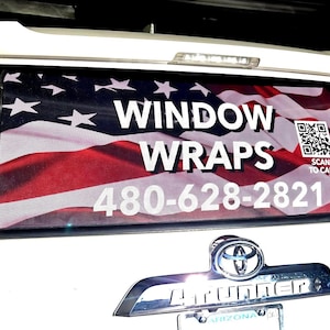 Op de afbeelding: Een witte SUV met een achterruitafbeelding met de Amerikaanse vlag. De woorden "WINDOW WRAPS" staan in witte letters, samen met een telefoonnummer en een QR-code met de tekst "SCAN TO CALL".