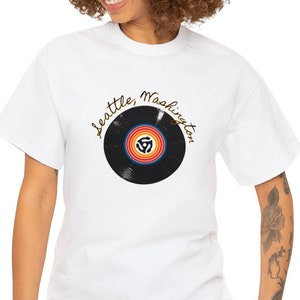 May include: White t-shirt featuring a black vinyl record graphic and the text "Seattle, Washington" in gold lettering.