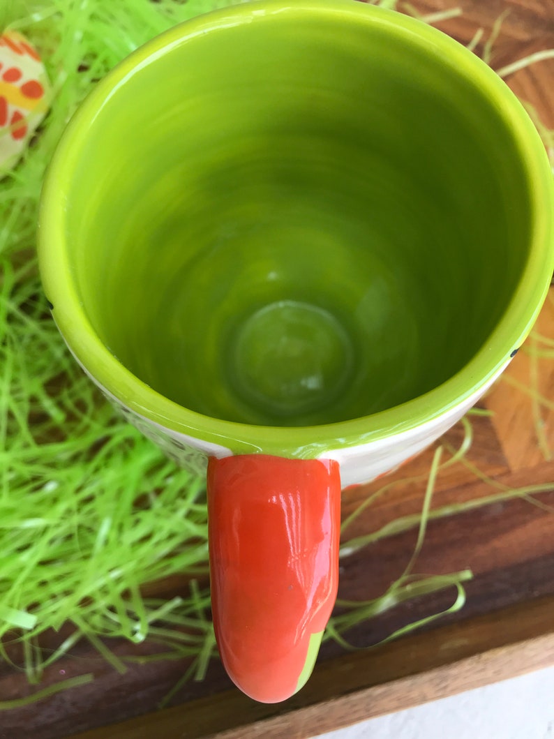 May include: A green ceramic mug with an orange handle. The mug is empty and has a glossy finish.