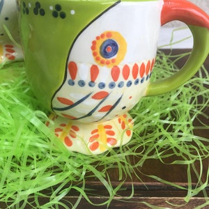 May include: A green and white ceramic mug with a colorful design. The mug has a handle and a wide base. The mug is sitting on a bed of green grass.