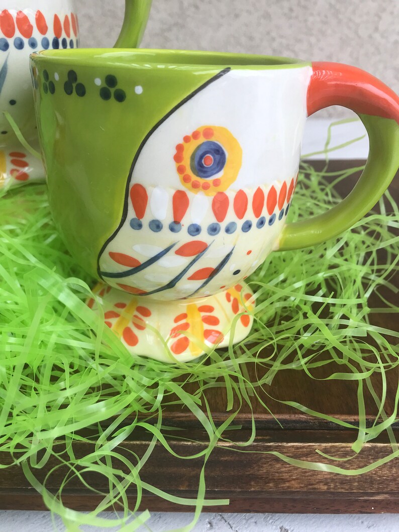 May include: A green and white ceramic mug with a colorful design. The mug has a handle and a wide base. The mug is sitting on a bed of green grass.