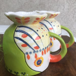 May include: Two green, white, orange, and blue ceramic mugs with a floral design. The mugs have a unique, wavy rim and a handle.