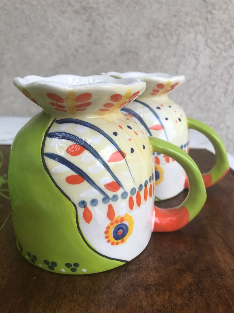 May include: Two green, white, orange, and blue ceramic mugs with a floral design. The mugs have a unique, wavy rim and a handle.