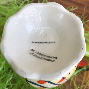 May include: White ceramic egg cup with a blue, yellow, and orange geometric design. The bottom of the cup reads "Dishwasher & Microwave Safe Made in China/Fabriqu&eacute; en Chine by Anthropologie".