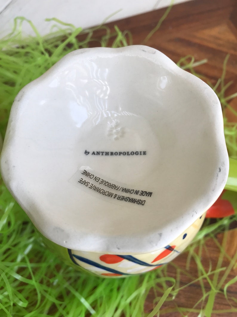 May include: White ceramic egg cup with a blue, yellow, and orange geometric design. The bottom of the cup reads "Dishwasher & Microwave Safe Made in China/Fabriqu&eacute; en Chine by Anthropologie".