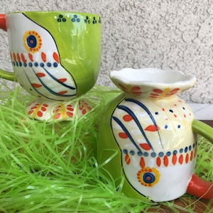 May include: Two ceramic mugs with colorful, abstract designs. The mugs have a green and white design with orange, blue, and yellow accents. The mugs are sitting on a bed of green grass.