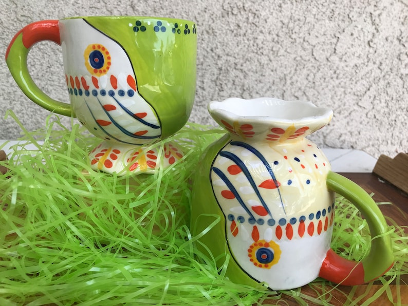 May include: Two ceramic mugs with colorful, abstract designs. The mugs have a green and white design with orange, blue, and yellow accents. The mugs are sitting on a bed of green grass.
