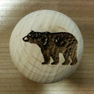 May include: A wooden knob with a detailed, brown bear design burned into the surface. The bear is depicted in profile, with intricate details. The knob is a light, natural wood color, and the bear image is centered on the round surface.