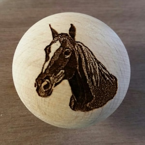 May include: A wooden sphere with a detailed, dark brown engraving of a horse's head. The horse has a white stripe down its face. The sphere is made of light-colored wood.