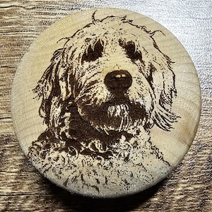 May include: A circular wooden disc featuring a dark, laser-engraved portrait of a shaggy-haired dog. The natural light-colored wood grain serves as the background for the detailed, high-contrast depiction of the canine's face and fur texture.