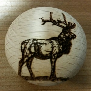 May include: A round, wooden cabinet knob with a detailed pyrography image of a standing elk. The elk is depicted in a side profile, with its antlers prominently displayed. The wood has a light, natural finish.