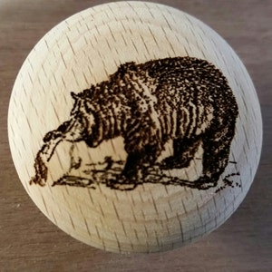 May include: A wooden ball with a detailed wood-burned image of a bear. The bear is depicted in a naturalistic style, with its fur and features carefully rendered. The ball is made of light-colored wood.