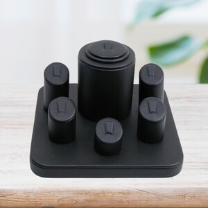 May include: Black 6-column ring display set with a central column and five smaller columns. The display is made of black velvet and has a rectangular base.