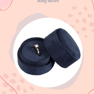 Könnte beinhalten: Eine dunkelblaue Samt-Ringbox mit Deckel. Die Box ist geöffnet und ein Diamantring befindet sich im Inneren. Der Ring ist in eine goldene Fassung gefasst.