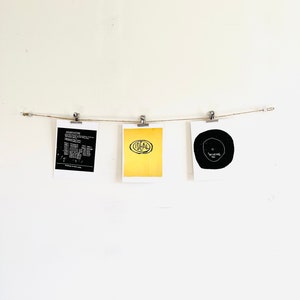 May include: Three black and white prints are hanging on a white wall with a string and clips. The prints are framed in white. The left print has text, the middle print has the word "TOTAL" in black on a yellow background, and the right print has a black circle with text inside.