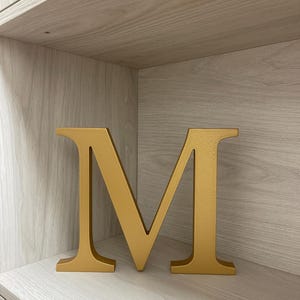 May include: A freestanding, three-dimensional capital letter M finished in a solid, matte gold color. The letter features a classic serif typeface with clean, straight lines and a flat base, allowing it to stand upright on a level surface.