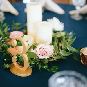 May include: A gold table number '3' sits on a table centerpiece with greenery, pink roses, and white candles. The table is covered in a dark blue tablecloth.