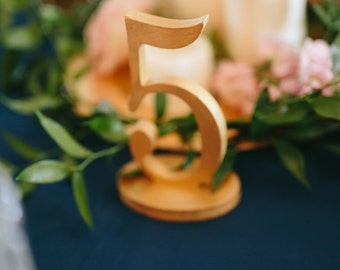 May include: A gold-colored wooden table number with the number "5" on it. The table number is sitting on a dark blue tablecloth. There are flowers and greenery in the background.