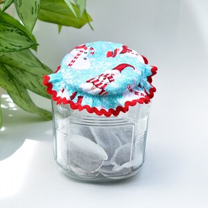 May include: A glass jar with a red and white scalloped fabric lid featuring a snowman and snowflake design. The jar is filled with tea bags.